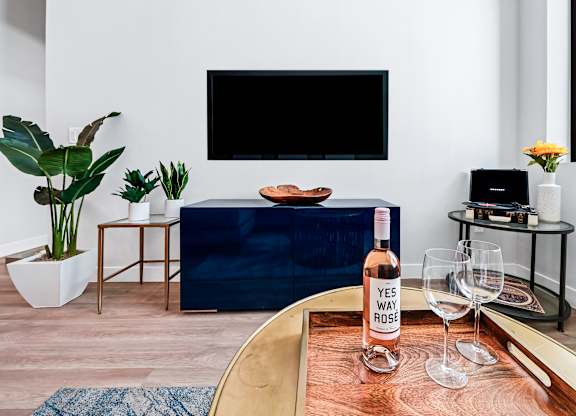 A living room with a blue couch, a wooden table with a bottle of wine and glasses, and a TV mounted on the wall.