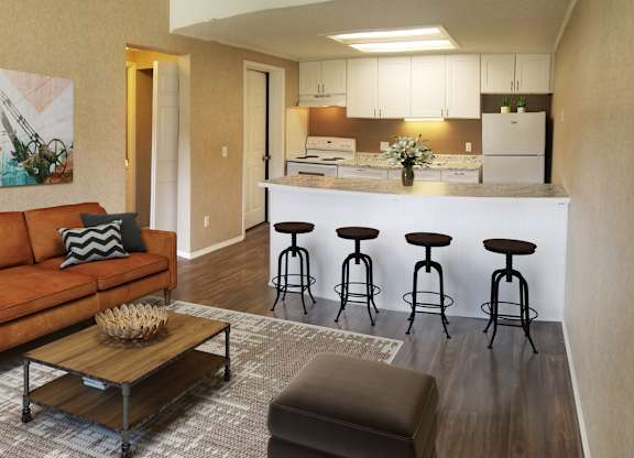 A kitchen with white cabinets and a refrigerator is adjacent to a living room with a brown couch and a coffee table.