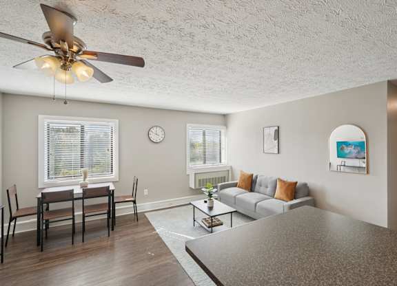 A living room with a grey couch, a dining table with chairs, a ceiling fan, and a clock on the wall.