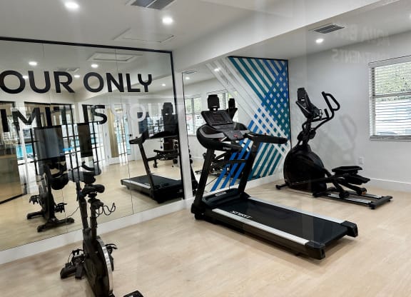 A gym with cardio equipment and a large window with your only limits signage on it at Aqua Bay Apartments in Naples, FL 34116