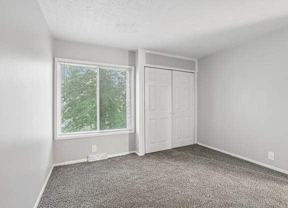 A room with a carpeted floor, a white door, and a window with a view of trees outside.