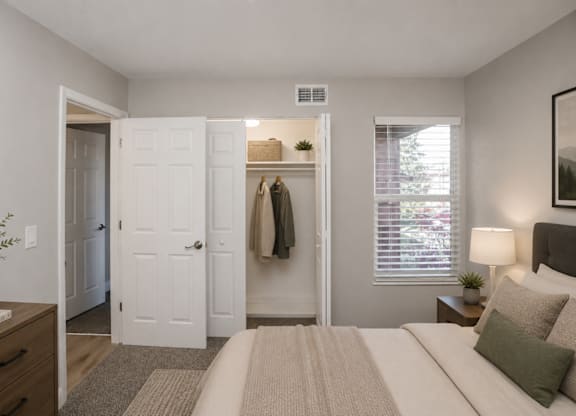 A bedroom with a bed, a nightstand, a dresser, a window with blinds, a coat rack with a coat hanging on it, and a plant on the dresser.
