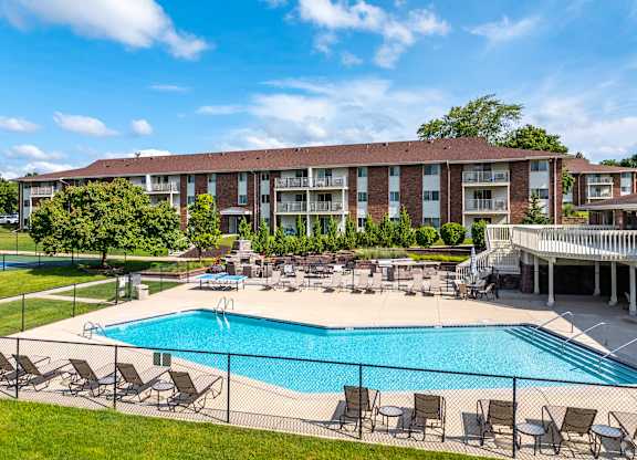 A large swimming pool surrounded by lawn chairs and umbrellas in front of a multi-story apartment building.