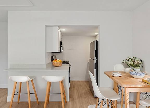 A kitchen with white walls and wooden floors, featuring a bar area with stools, a dining table with chairs, and a refrigerator.