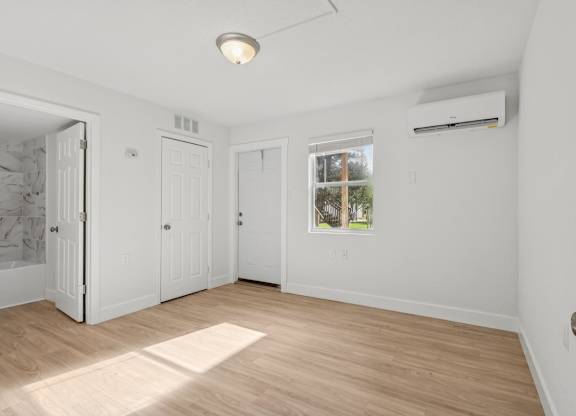 A room with white walls and wooden flooring, with a window showing a tree outside.