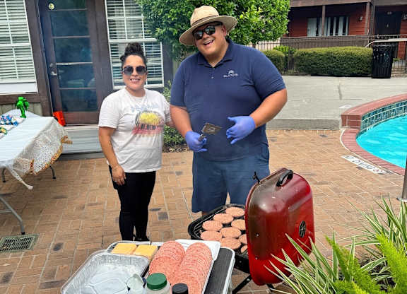 A man and a woman are standing by a table with food and a grill.