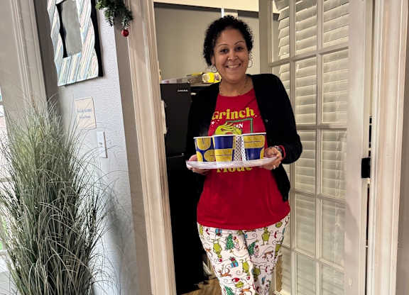 A woman in a red shirt and patterned pants holds a tray with a yellow box and a yellow cup.