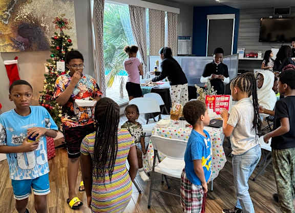 A group of children are gathered around a table with a Christmas tree in the background.