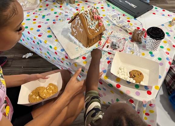A person is sitting at a table with a gingerbread house and cookies.