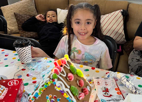 A young girl is sitting at a table with a gingerbread house in front of her.