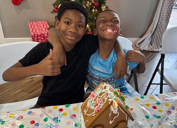 Two boys hugging and smiling in front of a gingerbread house.