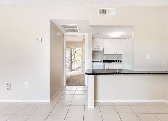 a living room and kitchen with a tiled floor, black countertops, white cabinets, and overhead lights