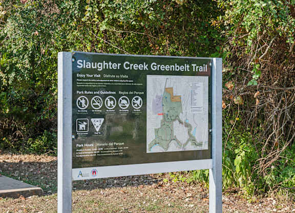 A sign for Slaughter Creek Greenbelt Trail is displayed in front of some trees.