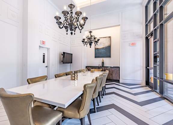 A long white table with chairs and a chandelier is in a room with a white wall.