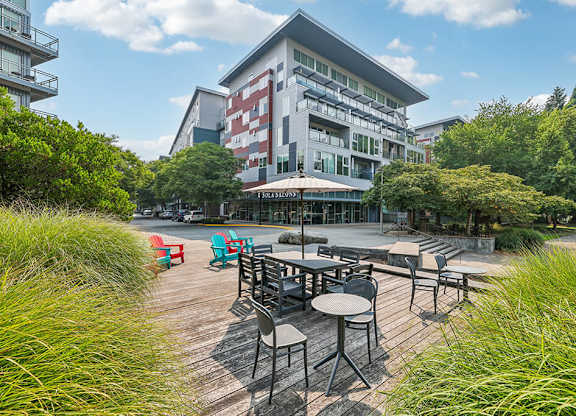 A wooden deck with chairs and tables is surrounded by greenery.