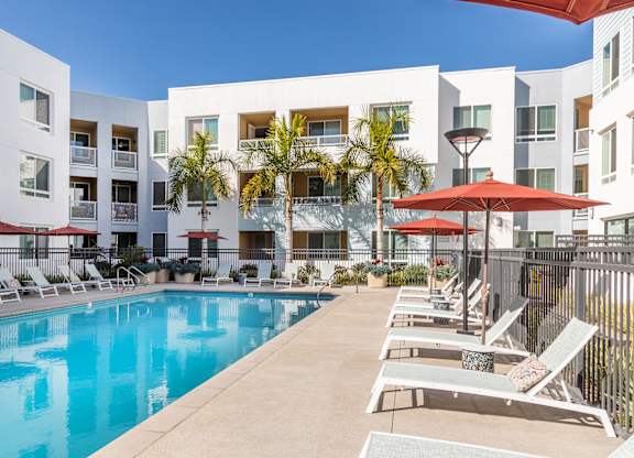 A swimming pool surrounded by lounge chairs and umbrellas in front of a white apartment building.