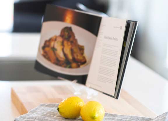 A book is open to a recipe for chicken, with two lemons on a cutting board beside it.
