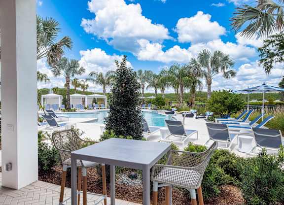 A table and chairs are set up on a patio with a pool and palm trees in the background.