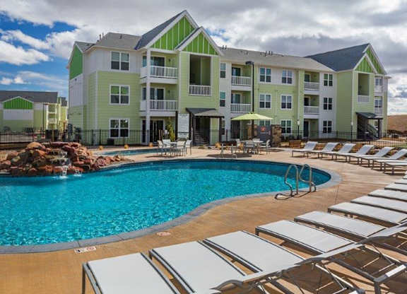 A large swimming pool surrounded by sun loungers in front of a multi-story apartment building.