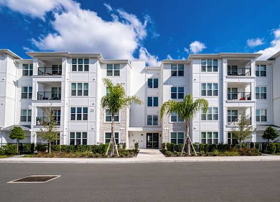 A row of white apartment buildings with balconies and trees in front.