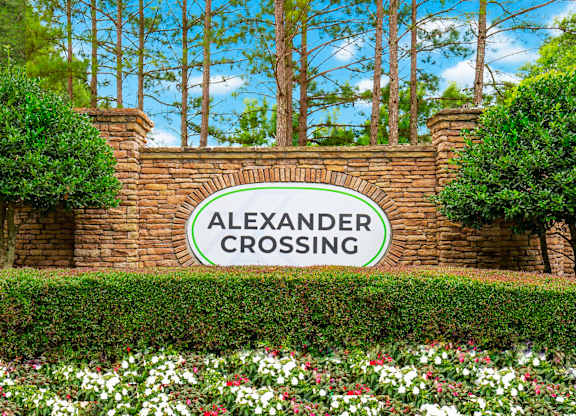 A sign that says Alexander Crossing is surrounded by greenery and flowers.