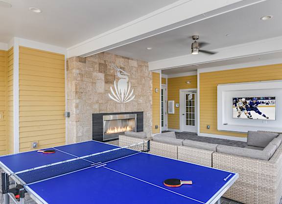 A blue ping pong table is in the middle of a living room with a fireplace and a painting on the wall.