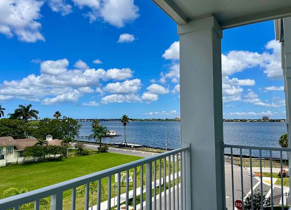 A balcony overlooks a body of water with a house and palm trees in the distance.