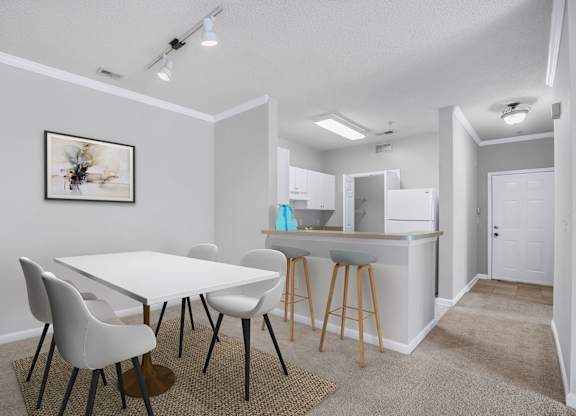 A white dining table with four chairs and a kitchen area in the background.