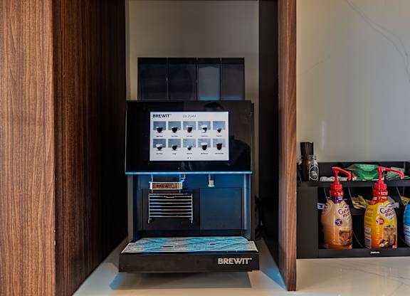 A Brewit coffee machine is on a counter with a shelf of drinks beside it.