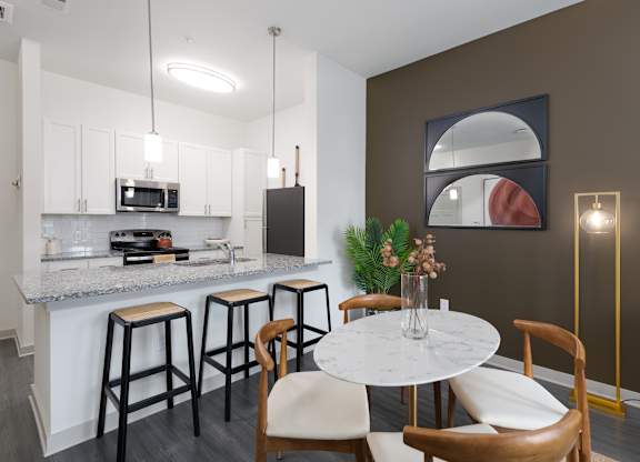 2-Bedroom Luxury Apartments in East Nashville, TN - Flats at Laurel and Pine - Dining Area with Vinyl Plank Flooring, Round Dining Table with Four Chairs, Floor Lamp, Breakfast Bar with Granite Countertop and Barstools, Pendant Lights, and View of Kitchen.