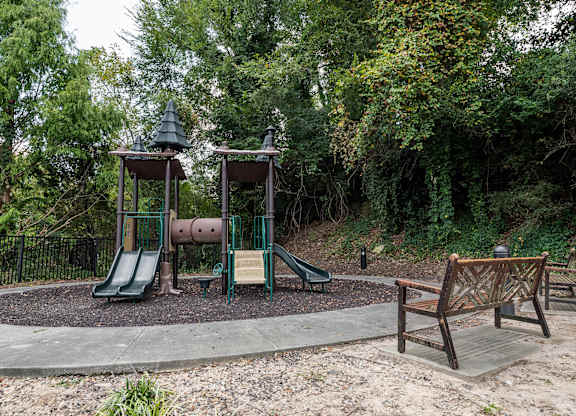 3-Bedroom Luxury Apartments in Atlanta, GA - The Eliza on Collier - Playground with a Multi-Play Structure on Wood Chips, Concrete Pathway, Wooden Bench, and Lush Green Trees.
