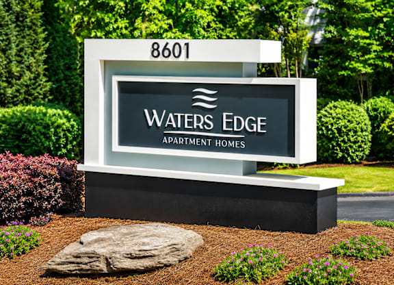 A sign for Waters Edge Apartment Homes is displayed in front of a green hedge.