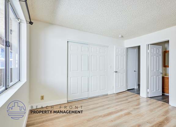 A room with white walls and wooden floors, featuring a white door and a window with a view of buildings outside.