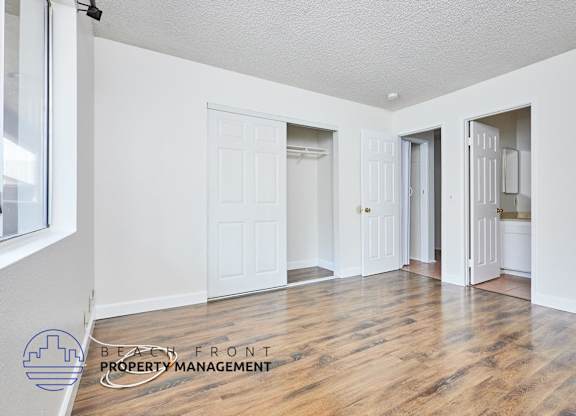 A spacious room with wooden floors and white walls, with a logo for Beach Front Property Management in the bottom left corner.