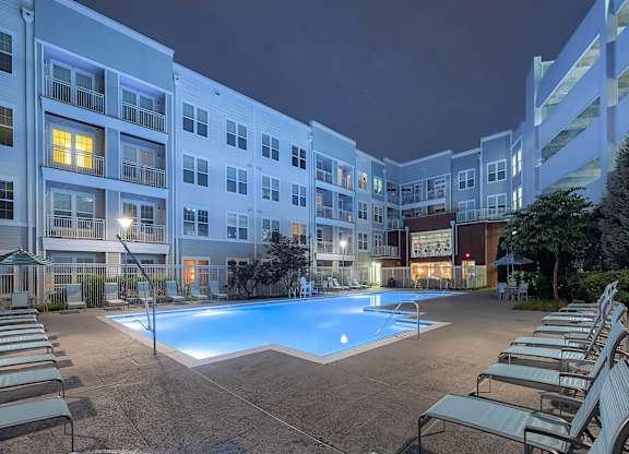 an outdoor swimming pool with chaise lounge chairs at night in front of an apartment building