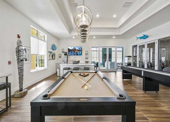 A pool table is in the middle of a room with a TV and a fish decoration on the wall and a catering kitchen is in the background..