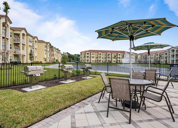 A patio with a table and chairs and umbrella next to BBQ grills in front of apartment buildings.