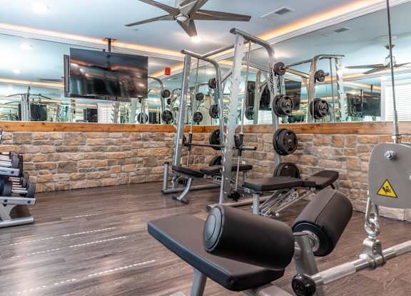 A gym with a variety of equipment including a bench press, a squat rack, and a television mounted on the wall at Mode at Melbourne, LLC Apartments, Florida