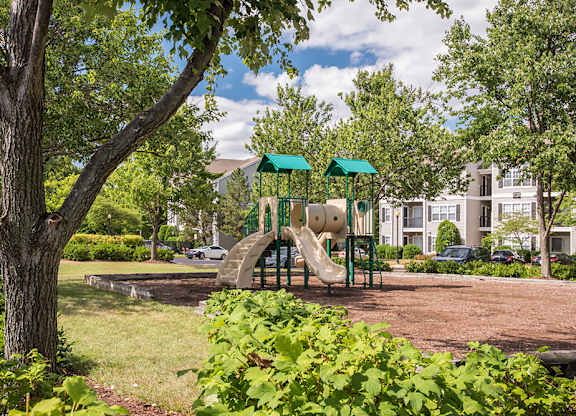 a wooded view of a playground with apartment buildings in the background.