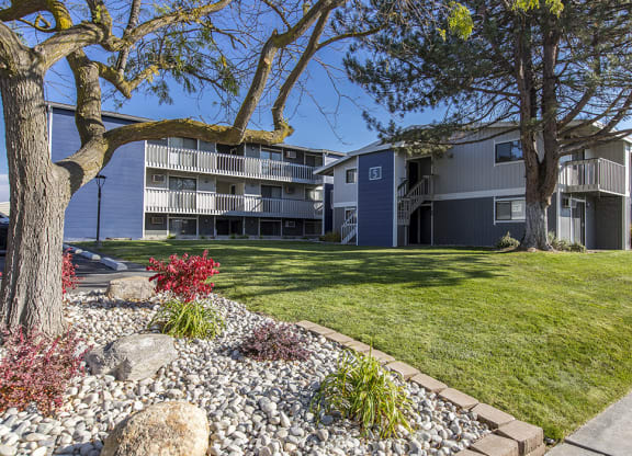 Exterior view of manicured grounds and building. Rock garden, large trees and blue skies in the background at The Lakes Apartments, Moses Lake, WA.