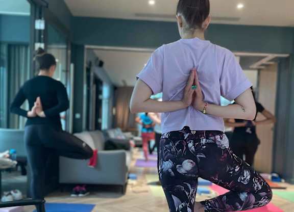 A woman in a purple shirt and floral pants is practicing yoga in a room with other people in the background. at Cherrywood Apartments, D18 VY3V