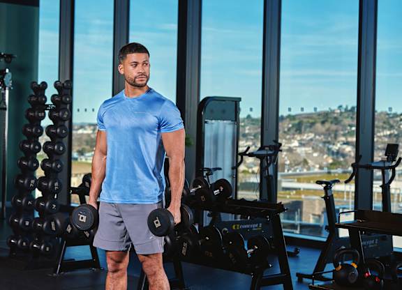 A man in a blue shirt and grey shorts is standing in a gym holding a dumbbell at Cherrywood Apartments, D18 VY3V, DUBLIN 18