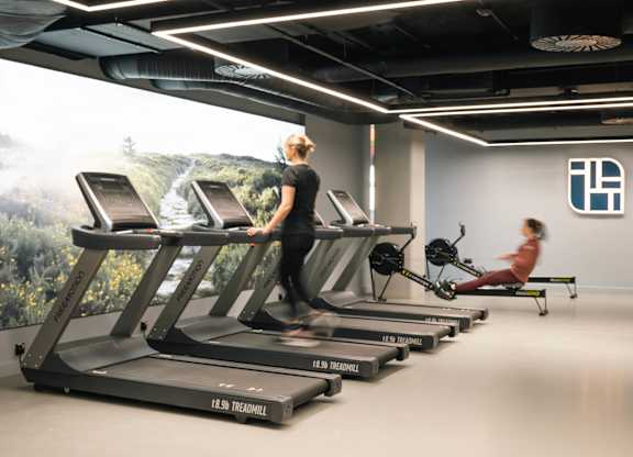A woman is running on a treadmill in a gym with a wall mural of a forest at Cherrywood Apartments, D18 VY3V