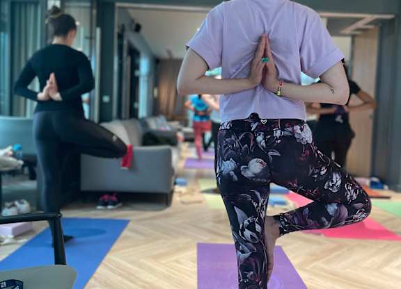 A woman in a purple shirt and floral pants is practicing yoga in a room with other people in the background at Cherrywood Apartments, D18 VY3V