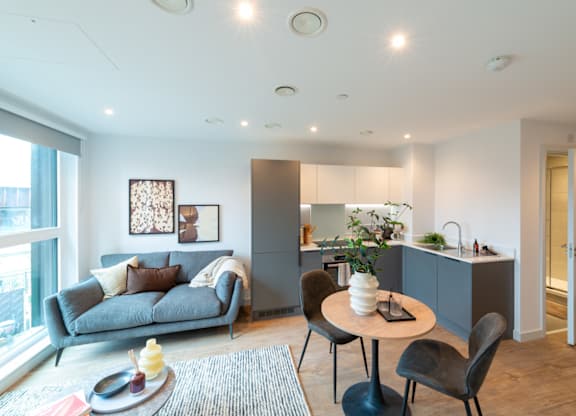 A modern living room with a grey couch, a round table with a vase of flowers, and a kitchen area in the background.