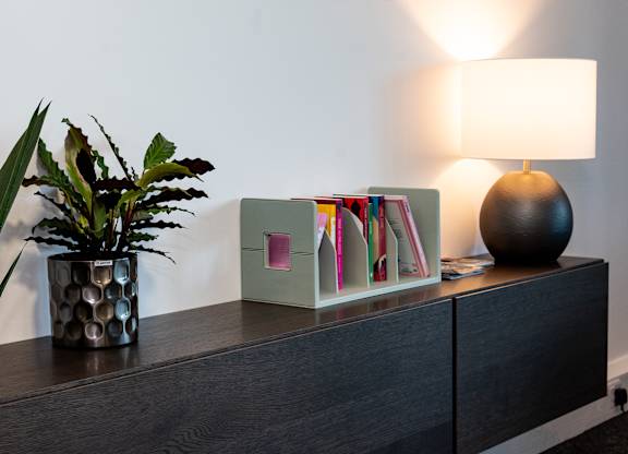A black vase with a green plant sits on a wooden dresser next to a lamp and a box of magazines.