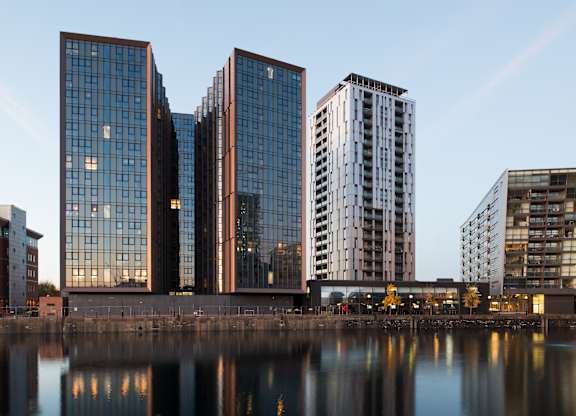 A group of modern buildings are reflected in the water in front of them.