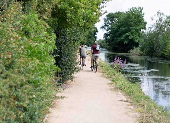 Two people riding bikes on a path next to a river at The Woodlands Apartments, Milton Keynes, MK5 6GS