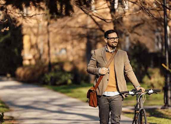 A man in a brown jacket and glasses is walking with a bicycle and holding a brown bag at Origins Apartments, Cambridge, CB3 0UX