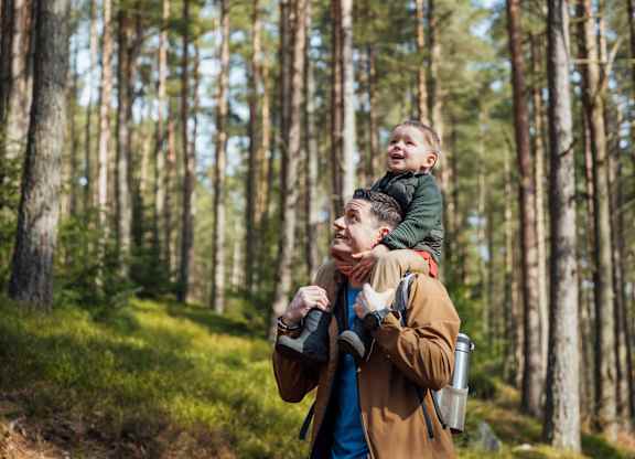 A man is carrying a child on his back while holding a camera in a forest at The Woodlands Apartments, Milton Keynes